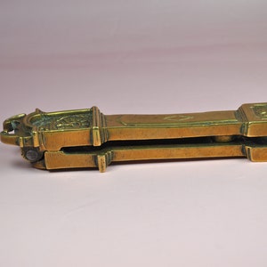 May include: A brass nutcracker featuring a long, rectangular body with a hinged mechanism. The surface displays a weathered, metallic gold finish with engraved geometric diamond patterns and a decorative clock-like face on the top end.