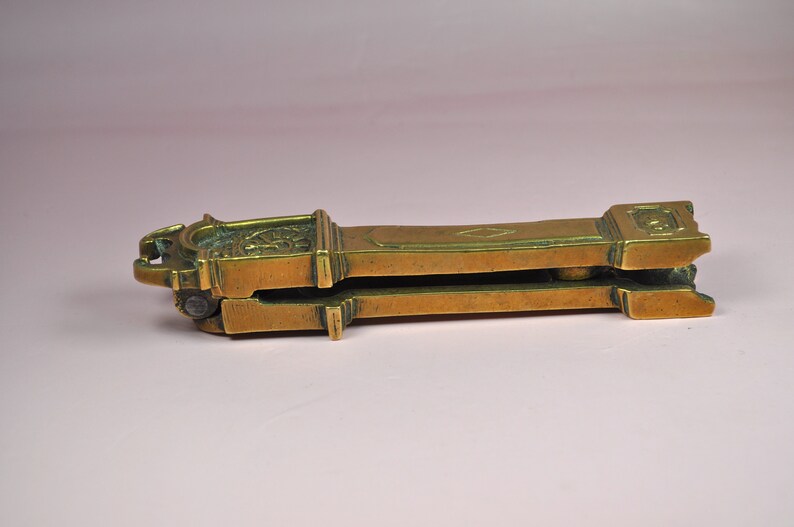 May include: A brass nutcracker featuring a long, rectangular body with a hinged mechanism. The surface displays a weathered, metallic gold finish with engraved geometric diamond patterns and a decorative clock-like face on the top end.