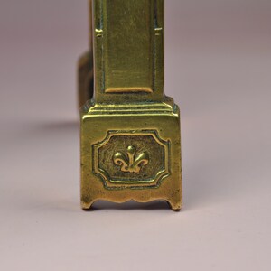May include: A brass furniture leg featuring a rectangular base with a raised fleur-de-lis emblem centered within a decorative border. The metal surface displays a warm, metallic gold tone with subtle signs of age and a smooth, polished finish.
