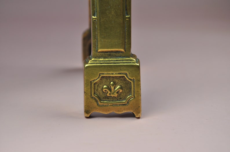May include: A brass furniture leg featuring a rectangular base with a raised fleur-de-lis emblem centered within a decorative border. The metal surface displays a warm, metallic gold tone with subtle signs of age and a smooth, polished finish.