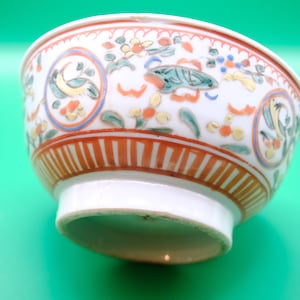 Puede incluir: Un cuenco de cerámica blanca con un patrón pintado a mano de motivos florales en naranja, verde y amarillo, junto con aves estilizadas dentro de marcos circulares. La base del cuenco está decorada con una serie de rayas verticales naranjas.