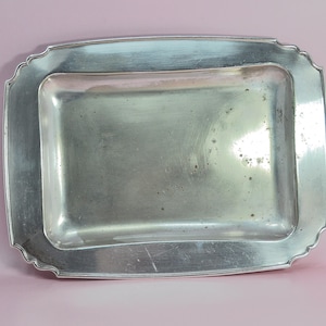 Puede incluir: Una bandeja rectangular plateada con un borde ligeramente festoneado. La bandeja tiene una superficie lisa y reflectante con pequeñas imperfecciones. El diseño es simple y elegante, adecuado para servir o exhibir.