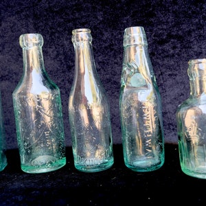 May include: Five antique glass bottles of different shapes and sizes, each with embossed text. The bottles are clear with a slight green tint, set against a dark background. The bottles appear to be vintage, possibly for medicine or other liquids.