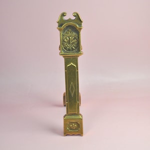 May include: A tall, narrow brass figurine shaped like a grandfather clock. The piece features an ornate top, a clock face with Roman numerals, a central panel with a diamond-shaped engraving, and a base decorated with a small fleur-de-lis motif.