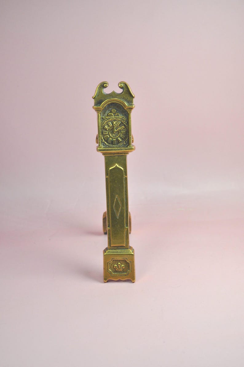 May include: A tall, narrow brass figurine shaped like a grandfather clock. The piece features an ornate top, a clock face with Roman numerals, a central panel with a diamond-shaped engraving, and a base decorated with a small fleur-de-lis motif.
