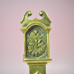 May include: A brass-colored metal object shaped like a miniature grandfather clock. The top features a decorative scroll design, while the clock face displays Roman numerals and two hands. The surface has a textured, aged finish with dark patina.