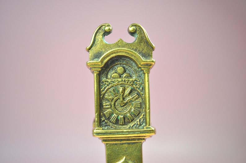 May include: A brass-colored metal object shaped like a miniature grandfather clock. The top features a decorative scroll design, while the clock face displays Roman numerals and two hands. The surface has a textured, aged finish with dark patina.