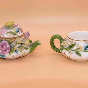 Può includere: Due contenitori in porcellana bianca a forma di teiere in miniatura, dotati di manici e beccucci verdi. I contenitori sono decorati con motivi floreali in rilievo dipinti a mano nei toni del rosa, giallo e blu, con dettagli dorati.
