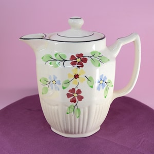 Op de afbeelding: Crèmekleurige keramische kan met deksel, handvat en tuit. Versierd met kleurrijke bloemmotieven in rood, geel en blauw, met groene bladeren. De basis heeft een verticaal geribbeld patroon. Een klassiek keukenartikel.