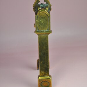 May include: A tall, narrow, metallic brass object with a weathered gold-toned finish. The structure features a rectangular base, a long vertical column, and a decorative top section with a curved arch and ornate, wing-like protrusions on each side.