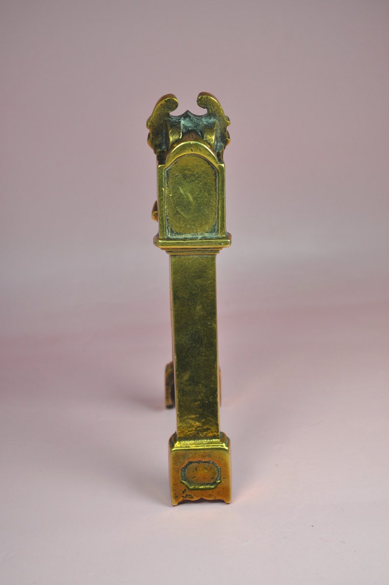 May include: A tall, narrow, metallic brass object with a weathered gold-toned finish. The structure features a rectangular base, a long vertical column, and a decorative top section with a curved arch and ornate, wing-like protrusions on each side.