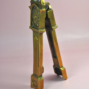 May include: A brass nutcracker shaped like a tall grandfather clock. The metal tool features a hinged design with a clock face detail at the top, a diamond-shaped engraving on the center panel, and a decorative base with a small floral motif.