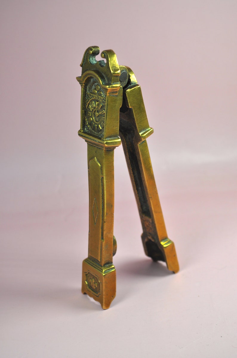 May include: A brass nutcracker shaped like a tall grandfather clock. The metal tool features a hinged design with a clock face detail at the top, a diamond-shaped engraving on the center panel, and a decorative base with a small floral motif.
