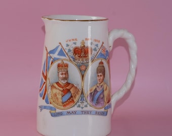Op de afbeelding: Witte keramische kan met een gouden rand en een gedraaide handgreep. De kan heeft portretten van een koning en koningin, vlaggen en de tekst "LONG MAY THEY REIGN". De datum "JUNE 26 1902" is ook zichtbaar.