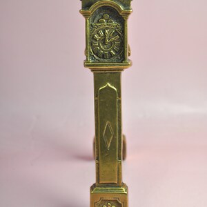 May include: A brass-colored metal figurine shaped like a tall grandfather clock. The piece features a detailed clock face with Roman numerals, a central diamond-shaped cutout on the long body, and a small fleur-de-lis emblem at the base.
