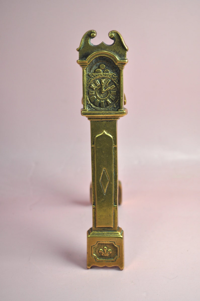 May include: A brass-colored metal figurine shaped like a tall grandfather clock. The piece features a detailed clock face with Roman numerals, a central diamond-shaped cutout on the long body, and a small fleur-de-lis emblem at the base.
