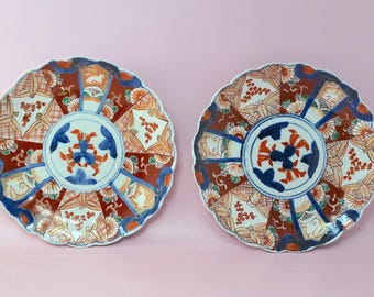 May include: Two round ceramic plates with scalloped edges, featuring a central blue and orange floral motif surrounded by alternating panels of deep red, white, and blue geometric patterns, floral sprigs, and stylized fan-like designs.