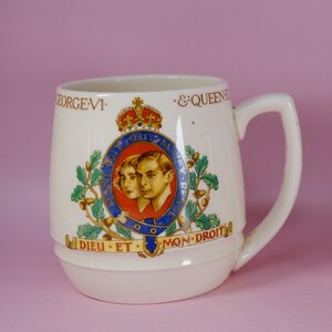 Può includere: Una tazza in ceramica color crema con un design commemorativo per Re Giorgio VI e la Regina Elisabetta. La parte anteriore mostra un ritratto centrale della coppia reale circondato da una giarrettiera blu, una corona, foglie di quercia e il testo.