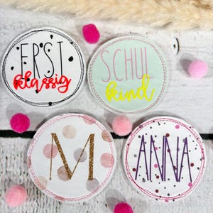 Könnte beinhalten: Vier runde Stoffuntersetzer mit gestickten Designs. Einer sagt "ERST Klassig" in roter Schrift, ein anderer sagt "SCHUL kind" in gelber Schrift, ein dritter hat ein goldenes "M" und der letzte sagt "ANNA" in Lila.