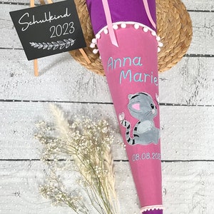 Könnte beinhalten: Ein rosa-lilafarbener Schultüte mit weißem Pomponbesatz. Die Tüte ist mit einer grauen Katze mit Schmetterling, dem Namen "Anna Marie" und dem Datum "08.08.202" verziert. Ein Schild aus Schiefertafel steht daneben und zeigt "Schulkind 2023".