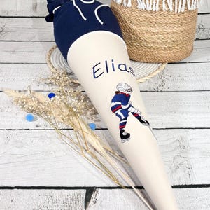 Könnte beinhalten: Eine cremefarbene Schultüte mit dem Namen "Elias" in Blau gestickt, zusammen mit einer Hockey-Spieler-Grafik. Die Oberseite ist marineblau. Ein geflochtener Korb mit weißem Fransenbesatz befindet sich im Hintergrund.