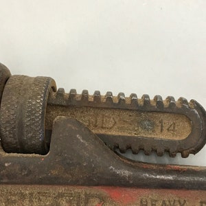 May include: Close-up of a well-worn, red and black pipe wrench. The wrench has a toothed jaw and a textured adjustment knob. The number "14" is visible on the jaw. The tool shows signs of age and use.