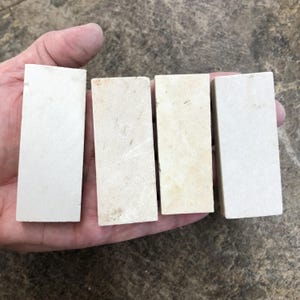 May include: Four rectangular, off-white sharpening stones of varying shades are held in a hand. The stones appear to be made of a porous material, with slight variations in color and texture. The stones are arranged side by side.