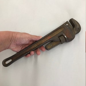 May include: A heavy-duty 35.5 cm pipe wrench with a rusty brown finish. The wrench has a Ridgid brand name and is held by a hand. The wrench is designed for gripping and turning pipes.