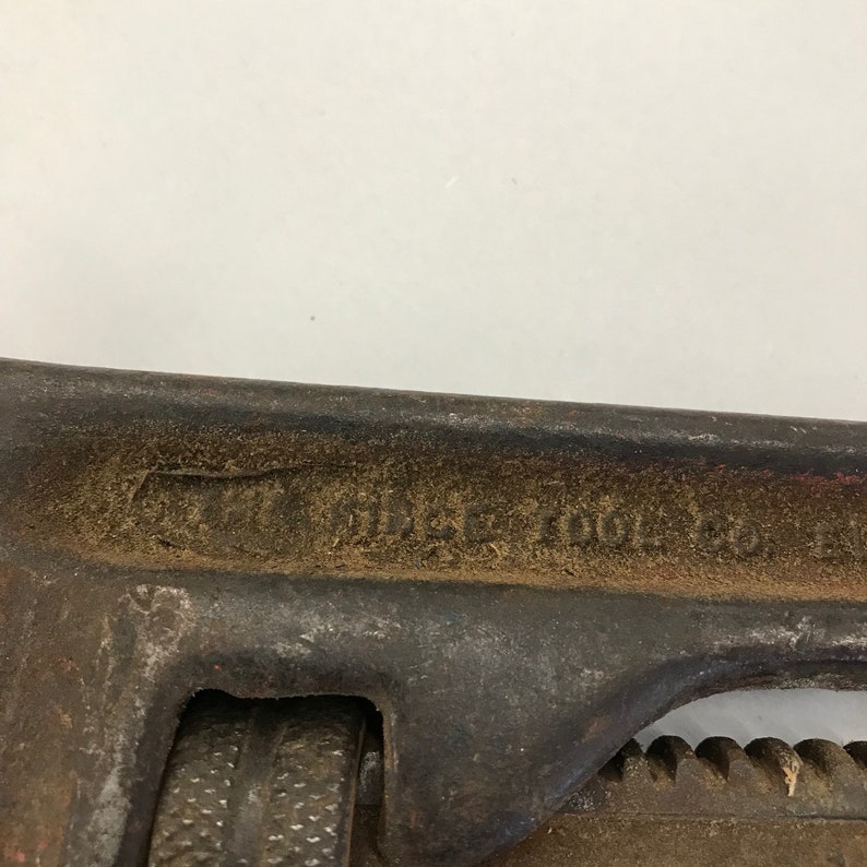May include: Close-up of a vintage RIDGID TOOL CO. pipe wrench. The wrench is dark brown with visible wear and tear, and the text "RIDGID TOOL CO." is embossed on the side. The adjustable jaw and handle are visible.