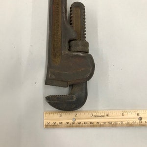 May include: A rusty, adjustable pipe wrench with a toothed jaw. The wrench is dark brown with red accents and shows signs of wear. A ruler is placed next to the wrench, measuring up to 15 cm.