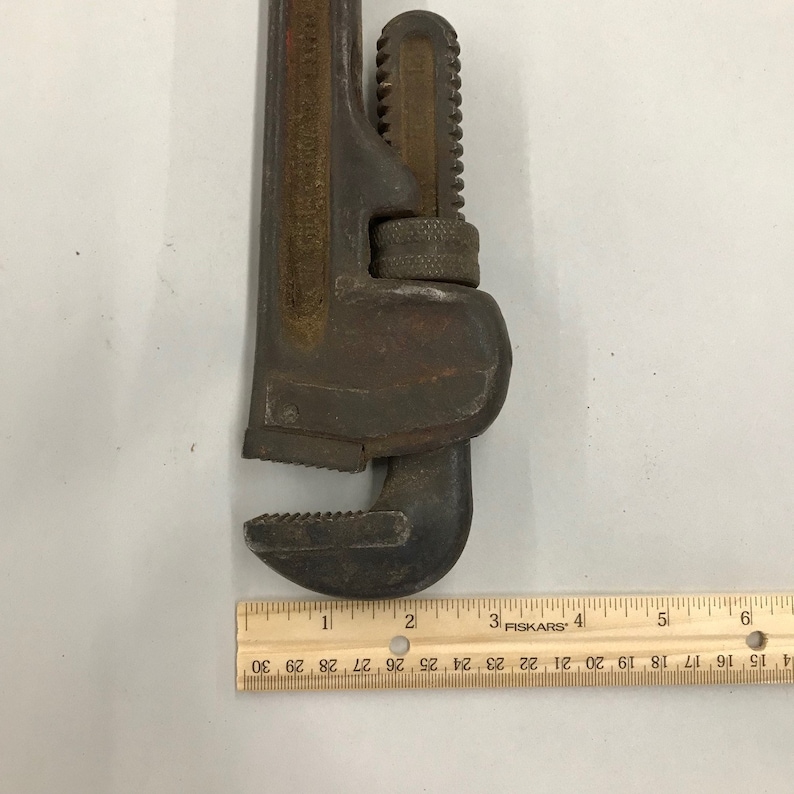 May include: A rusty, adjustable pipe wrench with a toothed jaw. The wrench is dark brown with red accents and shows signs of wear. A ruler is placed next to the wrench, measuring up to 15 cm.