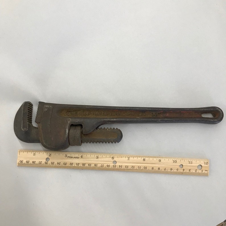 May include: A vintage pipe wrench with a dark, weathered appearance. The wrench has a long handle with a hole at the end and adjustable jaws. A ruler is placed next to the wrench for scale.