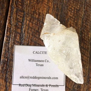 May include: A clear calcite crystal from Williamson Co., Texas. The crystal is on a white card with the text "CALCITE Williamson Co., Texas allen@reddogminerals.com Red Dog Minerals & Fossils Forney, Texas".