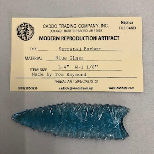 May include: A blue glass arrowhead replica, 4 inches long and 1 1/8 inches wide, made by Tom Raymond. The arrowhead is serrated and has a modern reproduction artifact label attached.