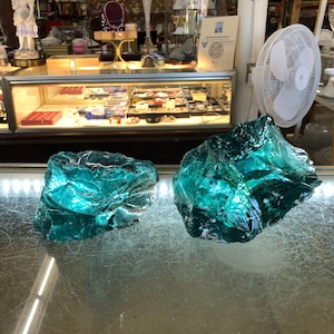 May include: Two large, irregular chunks of translucent teal-colored glass with jagged, reflective surfaces. The pieces feature deep blue-green hues and sharp, uneven edges, displaying a crystalline appearance with internal light refraction.