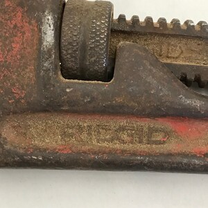 May include: Close-up of a vintage pipe wrench. The tool is primarily dark brown with red accents and shows signs of wear. The adjustable jaw and handle are visible, with the word "RIDGID" stamped on the side. A classic tool for plumbing and mechanical work.