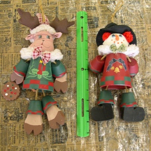 May include: Two Christmas ornaments, a reindeer and a snowman, both made of painted terracotta pots. The reindeer is brown and green with a red and white plaid scarf. The snowman is white with a black top hat, red mittens, and a black and white striped scarf. Both ornaments have bells hanging from them.