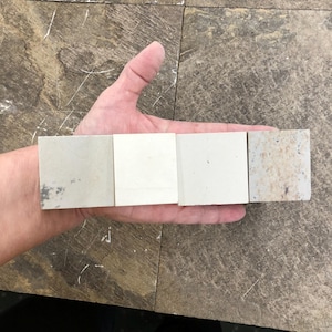Puede incluir: Una mano sosteniendo cuatro baldosas cuadradas con diferentes colores y texturas. Las baldosas varían de gris claro a blanco roto y beige moteado. Probablemente muestras para un proyecto de construcción o diseño.