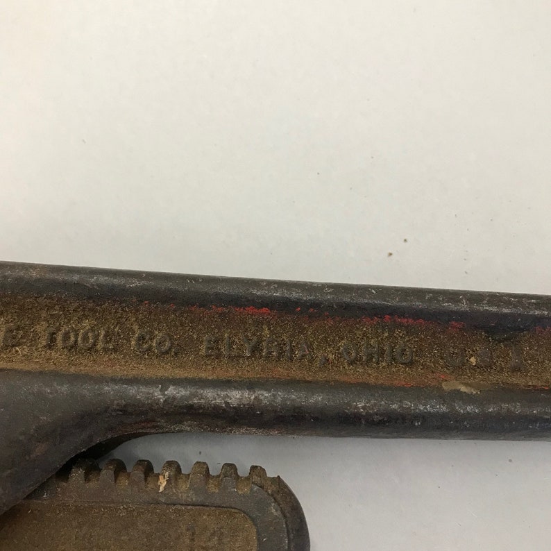 May include: A black and red pipe wrench with the text "ELYRIA, OHIO" visible. The wrench has a toothed jaw and a worn appearance, suggesting it is vintage or well-used. The tool is designed for gripping and turning pipes.