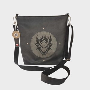 May include: Black crossbody bag with a dragon design. The bag features a circular emblem with a dragon's head in the center. It has a long, adjustable strap and a small tag attached. The bag is made of a dark, textured material.