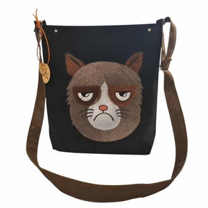 May include: A black canvas shoulder bag with a brown embroidered cat face. The cat has a grumpy expression with brown fur and white whiskers.