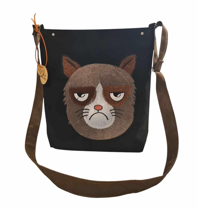 May include: A black canvas shoulder bag with a brown embroidered cat face. The cat has a grumpy expression with brown fur and white whiskers.