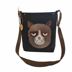 May include: Black canvas shoulder bag with a brown adjustable strap. The bag features an embroidered grumpy cat face.
