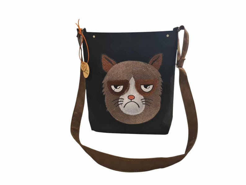 May include: Black canvas shoulder bag with a brown adjustable strap. The bag features an embroidered grumpy cat face.
