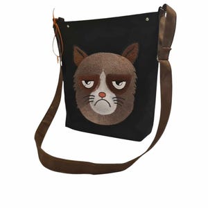 May include: A black canvas shoulder bag with a brown strap. The bag features an embroidered image of a grumpy cat with brown fur and white facial features.
