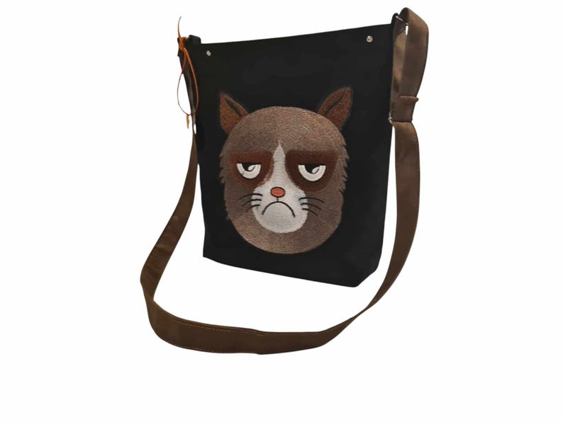 May include: A black canvas shoulder bag with a brown strap. The bag features an embroidered image of a grumpy cat with brown fur and white facial features.