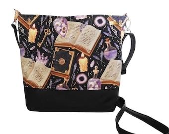 Pode incluir: Uma bolsa tote de tecido preto com uma estampa de livros, velas e frascos de poções em roxo, branco e dourado. A bolsa tem uma alça ajustável preta.