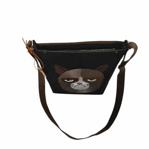 May include: A black canvas shoulder bag with a grumpy cat face embroidered on the front. The bag has a brown adjustable strap.