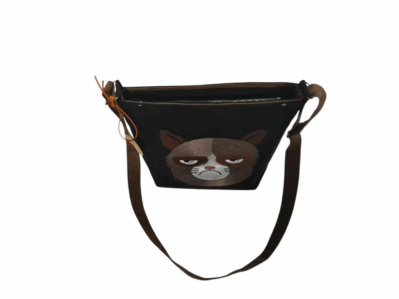 May include: A black canvas shoulder bag with a grumpy cat face embroidered on the front. The bag has a brown adjustable strap.