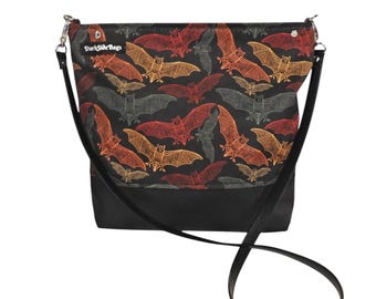 Pode incluir: Uma bolsa de ombro preta com uma alça longa ajustável. A bolsa tem um tecido preto com um padrão de morcegos laranja, vermelho e cinza.